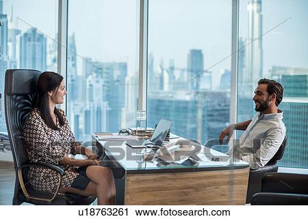 Stock Image - Businesswoman and man at desk with window view, Dubai, United Arab Emirates. Fotosearch