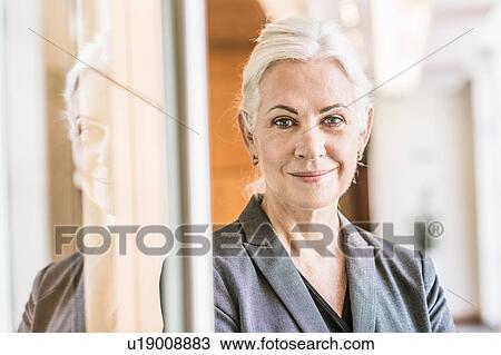 Stock Image - Businesswoman leaning against glass door looking at camera smiling. Fotosearch