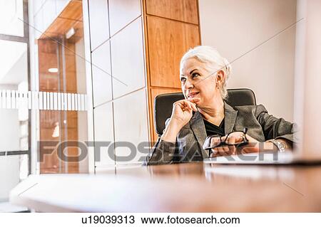Businesswoman sitting at desk holding glasses hand on chin looking away View Large Photo Image Stock Image - Businesswoman sitting at desk holding glasses hand on chin looking away. Fotosearch