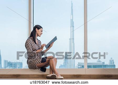 Stock Photo - Businesswoman using digital tablet sitting at window, Dubai, United Arab Emirates. Fotosearch