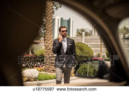 Stock Photo - Car window view of businessman talking on smartphone outside hotel, Dubai, United Arab Emirates. Fotosearch