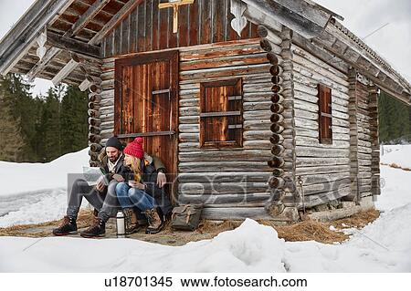 Stock Photography - Couple reading map sitting outside log cabin in winter, Elmau, Bavaria, Germany. Fotosearch
