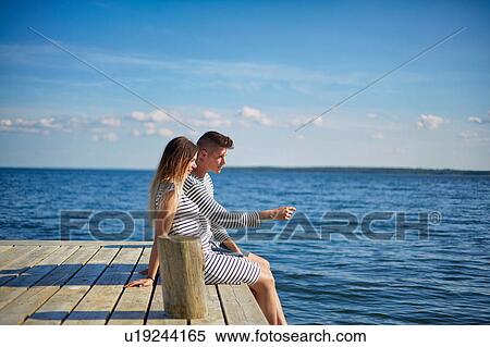 Stock Photography - Couple sitting on wooden pier, looking at view. Fotosearch