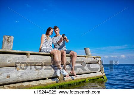 Stock Photography - Couple sitting on wooden pier, taking self portrait using smartphone. Fotosearch