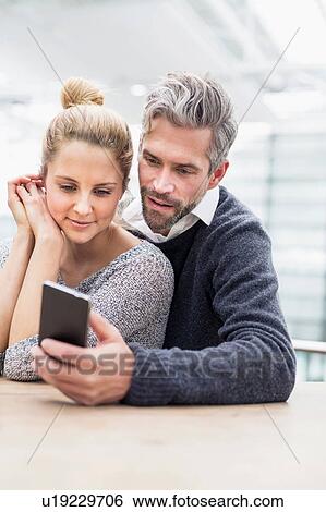 Couple sitting together at table, looking at smartphone View Large Photo Image Stock Photograph - Couple sitting together at table, looking at smartphone. Fotosearch