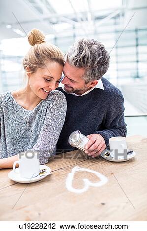 Stock Image - Couple sitting together, drinking coffee, heart shape made from sugar on table in front of them. Fotosearch