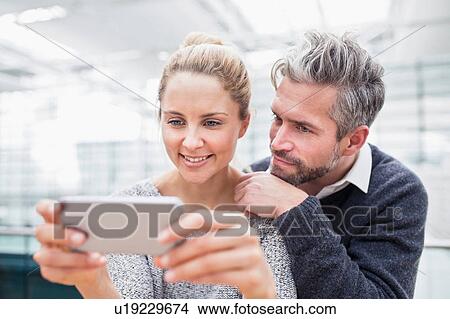 Couple sitting together, looking at smartphone View Large Photo Image Picture - Couple sitting together, looking at smartphone. Fotosearch