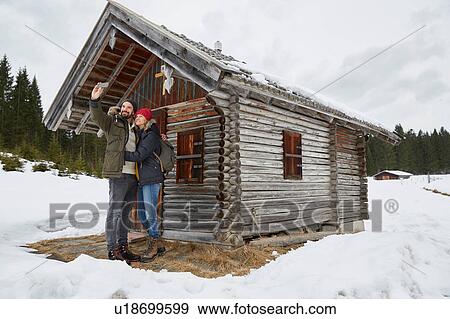 Stock Photo - Couple taking smartphone selfie outside log cabin in winter, Elmau, Bavaria, Germany. Fotosearch