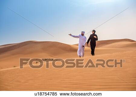 Stock Photography - Couple wearing traditional middle eastern clothes pointing from desert dune, Dubai, United Arab Emirates. Fotosearch