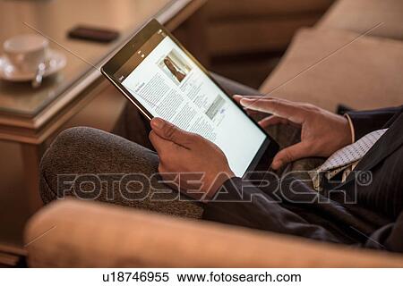 Stock Photography - Cropped shot of young businessman sitting on hotel sofa reading digital tablet. Fotosearch