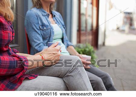 Stock Photograph - Cropped view of women sitting on chairs in front of shop holding coffee cups. Fotosearch