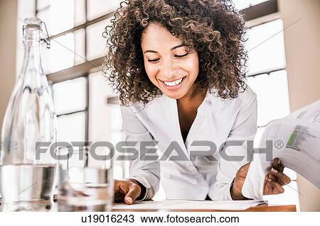 Stock Image - Curly haired business woman looking down at paperwork smiling. Fotosearch