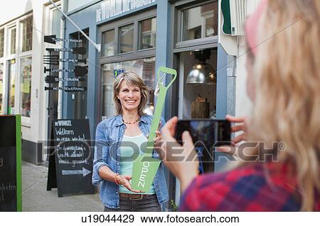 Stock Photo - Friend photographing woman in front of shop holding open sign. Fotosearch