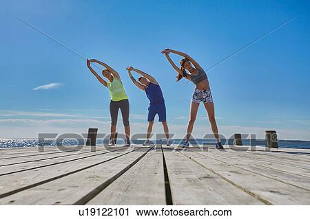 Stock Image - Friends on pier side by side arms raised bending over sideways stretching. Fotosearch