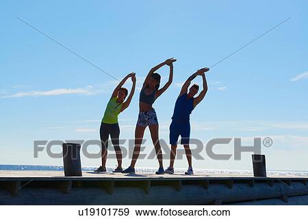 Friends on pier wearing exercise clothes arms raised, bending over sideways stretching View Large Photo Image Stock Photo - Friends on pier wearing exercise clothes arms raised, bending over sideways stretching. Fotosearch