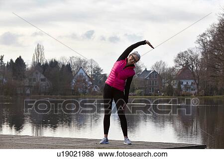 Stock Photo - Full length front view of woman in front of pond, arms raised bending over stretching. Fotosearch