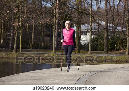 Stock Photograph - Full length front view of woman nordic walking by pond. Fotosearch