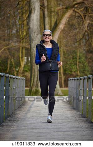 Stock Image - Full length front view of woman on wooden path jogging, looking at camera smiling. Fotosearch