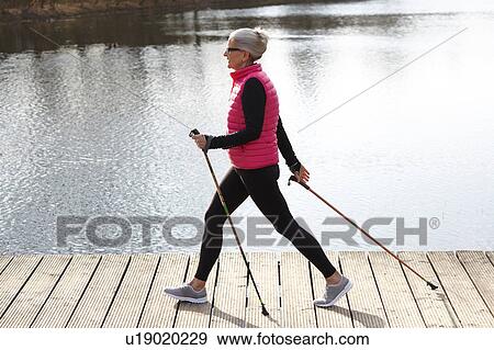 Stock Photo - Full length side view of woman nordic walking by pond. Fotosearch