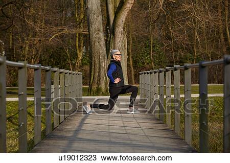 Stock Image - Full length side view of woman on wooden path, hands on hips lunging. Fotosearch