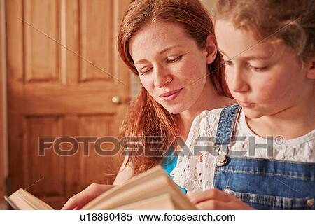 Stock Photography - Girl reading book with mother in living room. Fotosearch