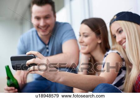 Stock Photo - Group of friends, sitting outdoors, drinking bottled beer, taking self portrait, using smartphone. Fotosearch