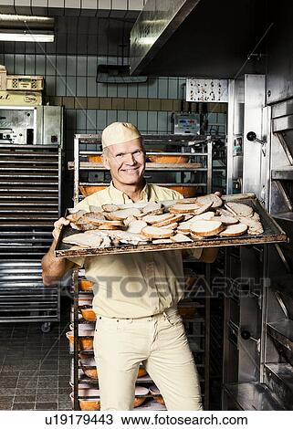 Stock Image - Happy baker placing tray of sliced bread into oven. Fotosearch