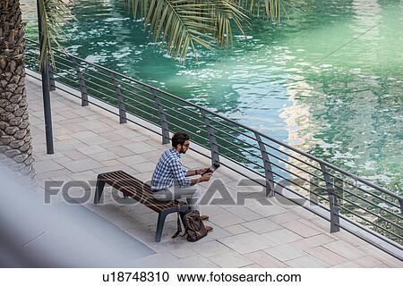 Stock Image - High angle view of young man sitting at waterfront using digital tablet, Dubai, United Arab Emirates. Fotosearch