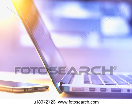 Stock Image - Laptop computer and mobile phone at desk with sunlight coming through window. Fotosearch