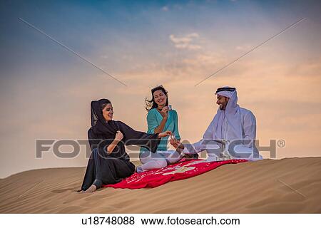 Stock Photo - Local couple wearing traditional clothes picnicing on desert dune with female tourist, Dubai, United Arab Emirates. Fotosearch