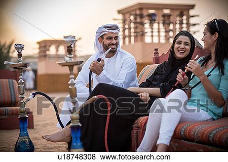 Local couple wearing traditional clothes smoking shisha on sofa with female tourist, Dubai, United Arab Emirates View Large Photo Image Stock Photo - Local couple wearing traditional clothes smoking shisha on sofa with female tourist, Dubai, United Arab Emirates. Fotosearch