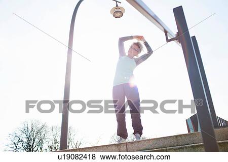 Stock Photo - Low angle view of woman on steps arms raised bending over sideways stretching. Fotosearch