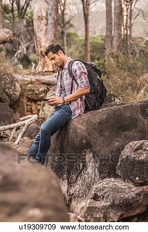 Stock Photo - Male hiker reading smartphone texts on forest rock formation, Deer Park, Cape Town, South Africa. Fotosearch