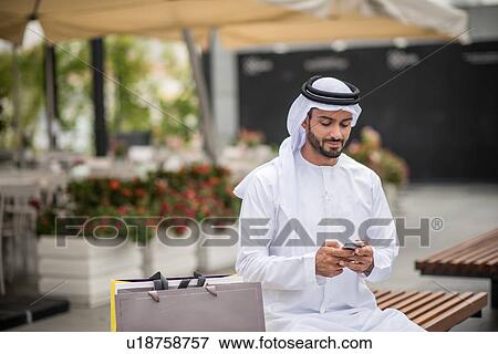 Male shopper wearing traditional middle eastern clothing sitting on bench reading smartphone text, Dubai, United Arab Emirates View Large Photo Image Stock Photo - Male shopper wearing traditional middle eastern clothing sitting on bench reading smartphone text, Dubai, United Arab Emirates. Fotosearch