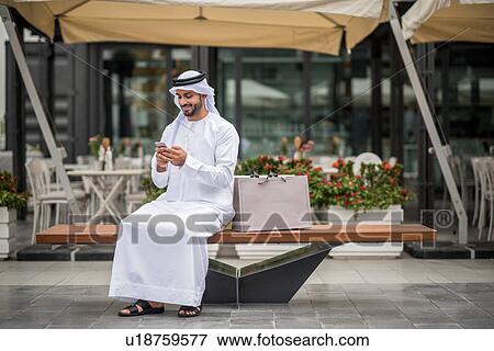 Male shopper wearing traditional middle eastern clothing sitting on bench reading smartphone text, Dubai, United Arab Emirates View Large Photo Image Stock Photo - Male shopper wearing traditional middle eastern clothing sitting on bench reading smartphone text, Dubai, United Arab Emirates. Fotosearch