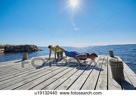 Stock Image - Man on pier doing press ups, legs raised on friend. Fotosearch