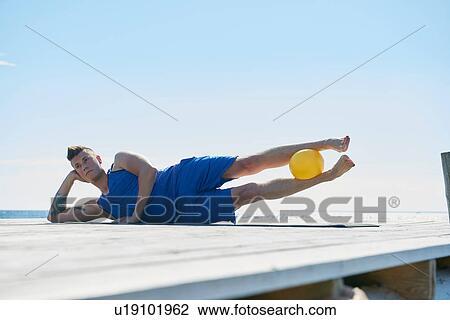 Stock Image - Man on pier lying on side resting on elbow, ball between legs. Fotosearch