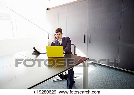 Stock Photo - Man sitting at desk using laptop making telephone call. Fotosearch