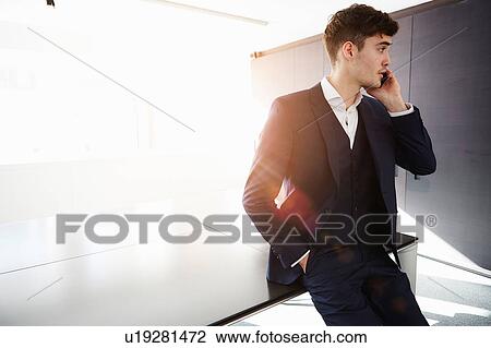 Stock Image - Man sitting on desk making telephone call looking away. Fotosearch