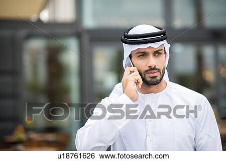 Man wearing dishdasha walking along street talking on smartphone, Dubai, United Arab Emirates View Large Photo Image Stock Photograph - Man wearing dishdasha walking along street talking on smartphone, Dubai, United Arab Emirates. Fotosearch