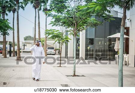 Man wearing traditional middle eastern clothing walking along street talking on smartphone, Dubai, United Arab Emirates View Large Photo Image Stock Image - Man wearing traditional middle eastern clothing walking along street talking on smartphone, Dubai, United Arab Emirates. Fotosearch