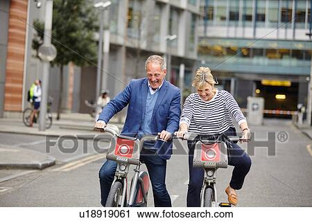 Stock Image - Mature dating couple laughing whilst cycling on hire bicycles, London, UK. Fotosearch
