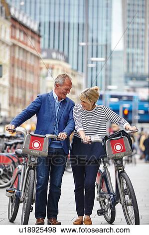Stock Photo - Mature dating couple laughing whilst pushing hire bicycles, London, UK. Fotosearch