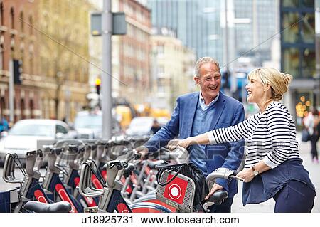 Stock Image - Mature dating couple removing hire bicycles from rack, London, UK. Fotosearch
