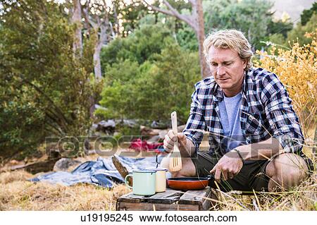 Stock Photography - Mature man frying breakfast on camping stove in forest, Deer Park, Cape Town, South Africa. Fotosearch