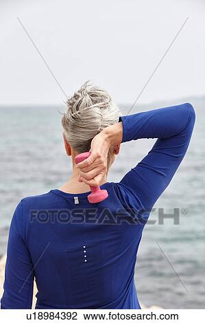 Stock Image - Mature woman beside sea, exercising with hand weights, rear view. Fotosearch