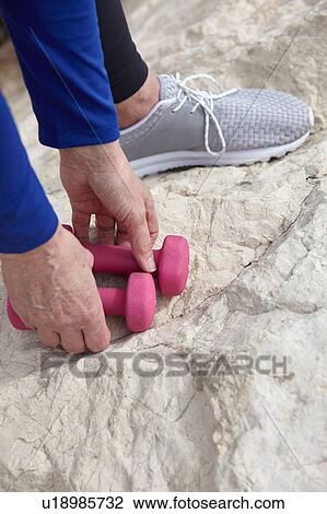 Stock Image - Mature woman on beach, picking up hand weights, low section. Fotosearch