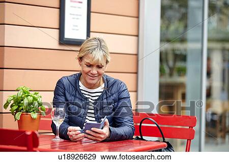 Stock Photo - Mature woman reading smartphone texts at sidewalk cafe. Fotosearch
