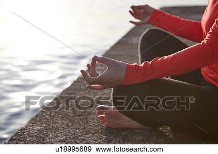 Stock Photo - Mature woman sitting beside water, in yoga position, low section. Fotosearch