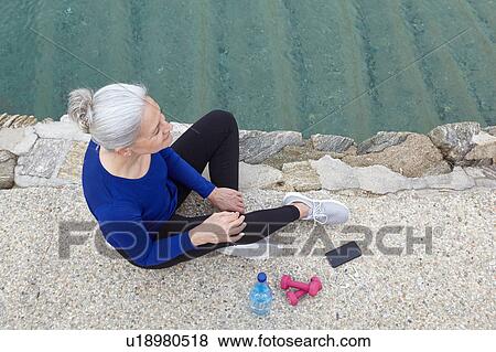 Stock Photo - Mature woman sitting outdoors, hand weights, water and smartphone beside her, elevated view. Fotosearch